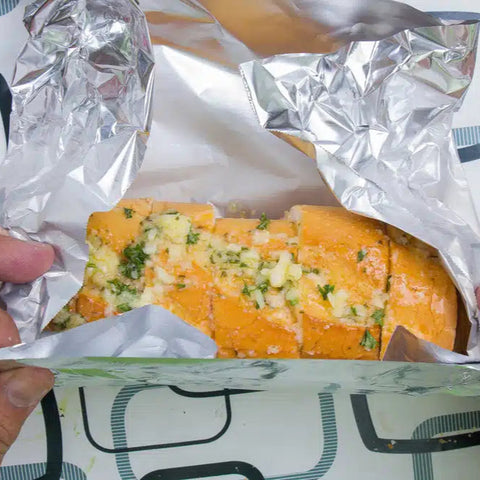 Person holding a piece of bread with cheese and herbs wrapped in aluminum foil.