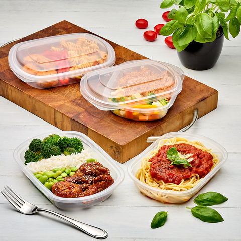 Several clear plastic meal prep containers filled with prepared food are arranged on a wooden cutting board and a light wood tabletop, with forks, basil leaves, and a small potted plant around them.