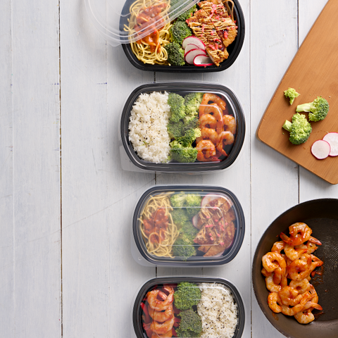 Four black plastic meal prep containers with clear lids are arranged vertically on a white wood plank surface, filled with different lunches including noodles, broccoli, shrimp, and rice. A wooden cutting board and a bowl of cooked shrimp are placed to the right as styling props.