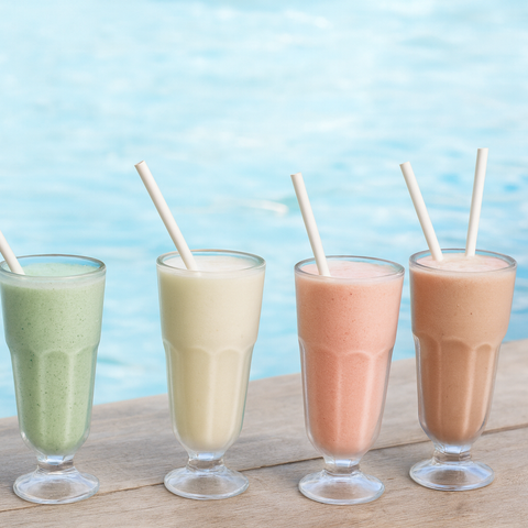 Four glasses of different colored smoothies with straws on a wooden surface against a blue sky.
